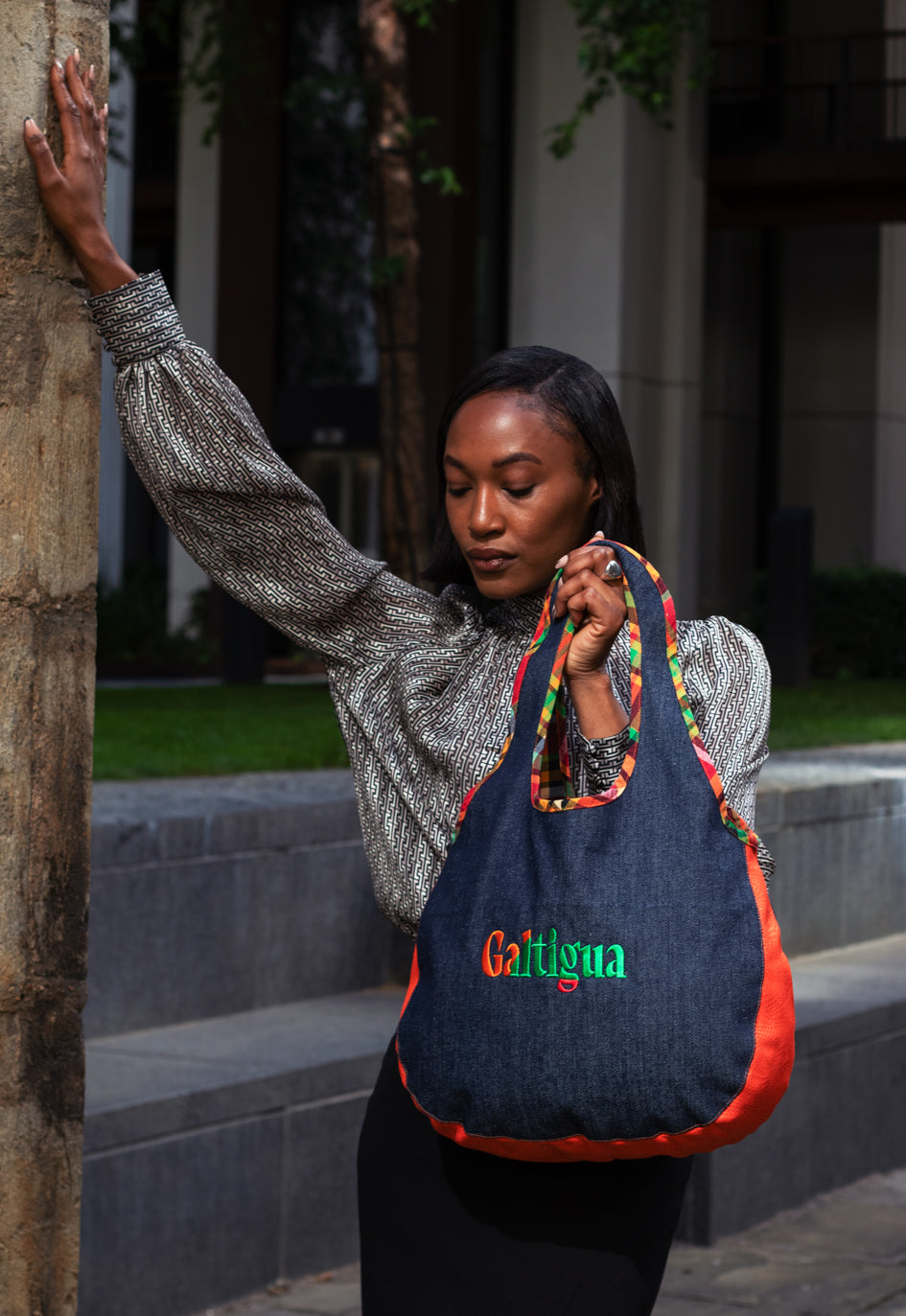 A young woman standing on a sidewalk leaning up against a post, holding a denim Hobo shoulder bag with 'Galtigua' text logo on it.