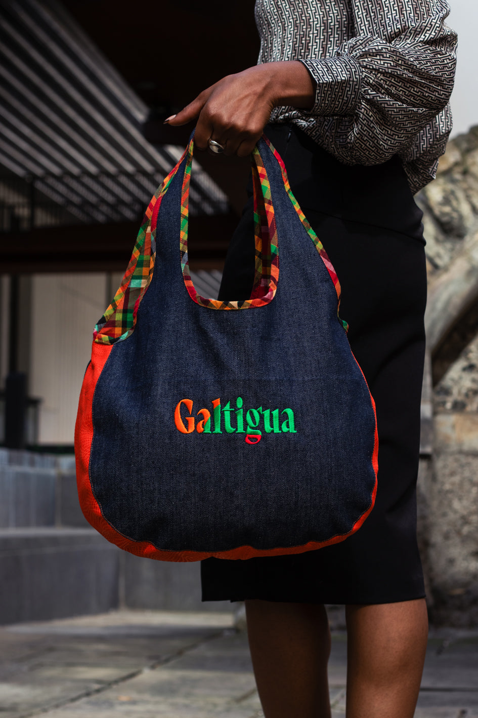 A young woman standing on a sidewalk, dressed formally holding a denim Hobo shoulder bag with 'Galtigua' text logo on it.