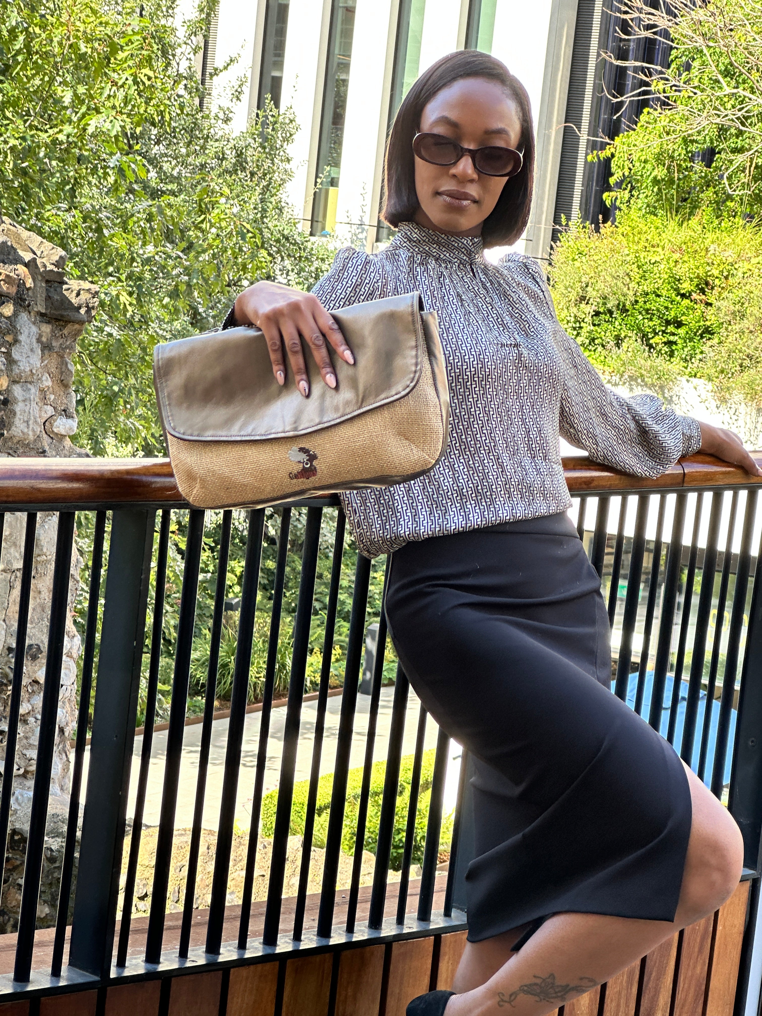 A young woman wearing a light coloured blouse, holding in her right hand a  Mahalia clutch bag with a Hessian fabric, bronze sheen leather, standing  against railings. 