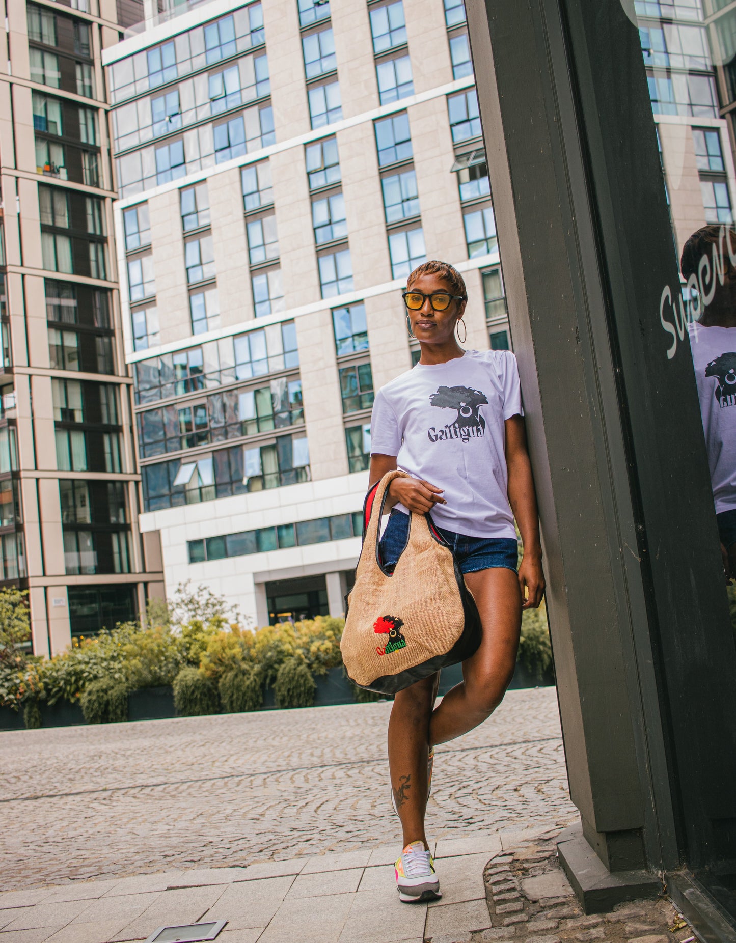 Young lady in a white Galtigua logo t-shirt and denim shorts, holding the Kalia Hobo style tote bag. She is leaning against the wall of a shop surrounded by office buildings.