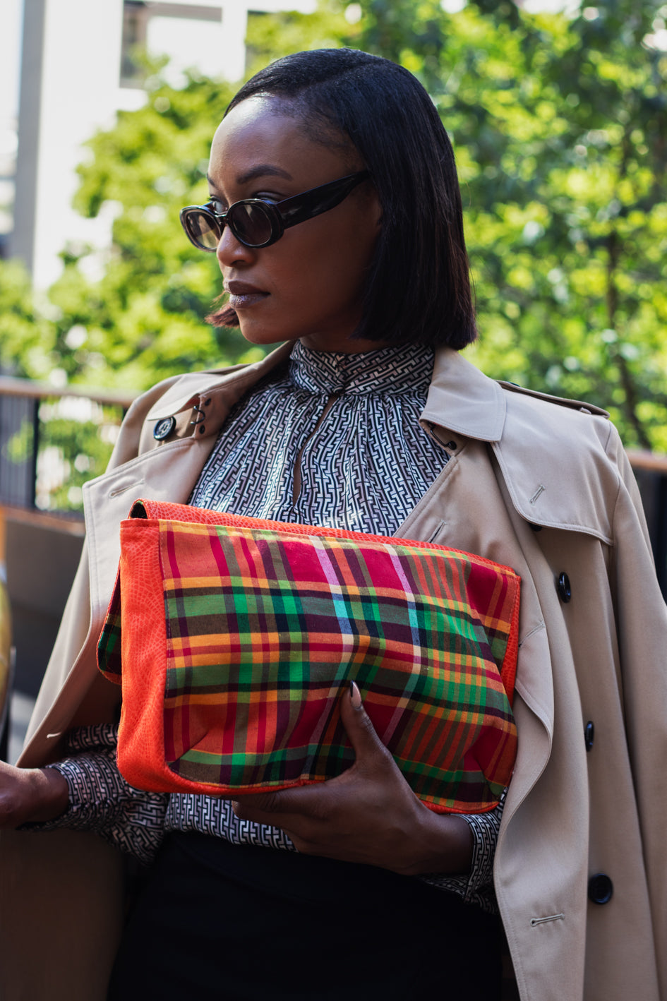 Young woman wearing sunglasses and a Mac over hers shoulders holding the Mahalia Papaya Orange Madras Print Clutch bag, against an office block & trees.