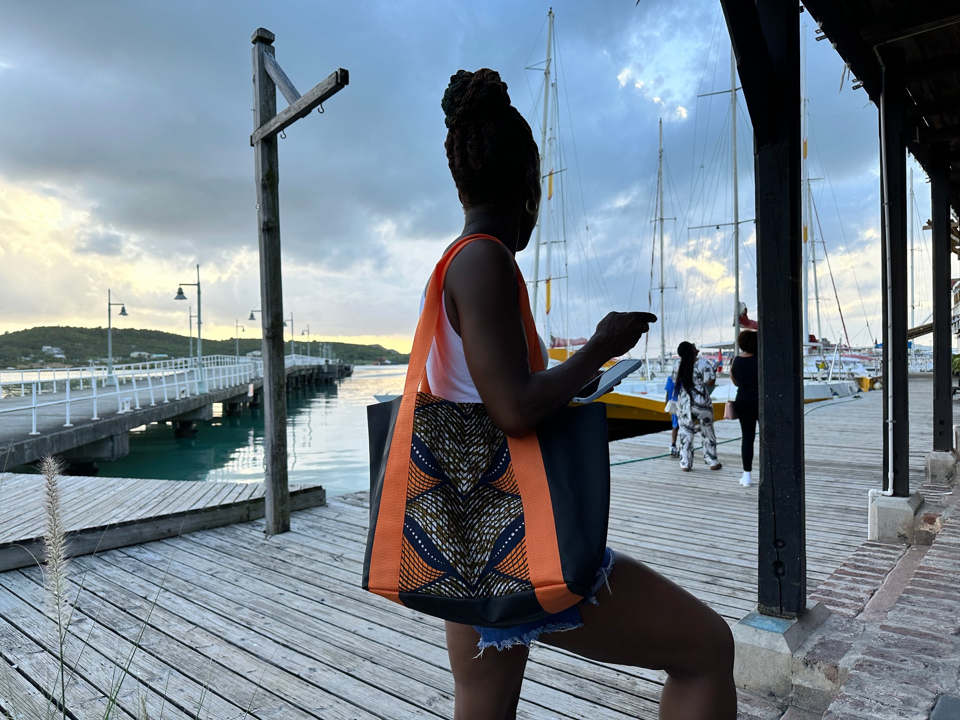 Woman dressed in jeans and a white vest top holding the Mini Kelaya over her right shoulder on the broad walk of wharf with docked boats.