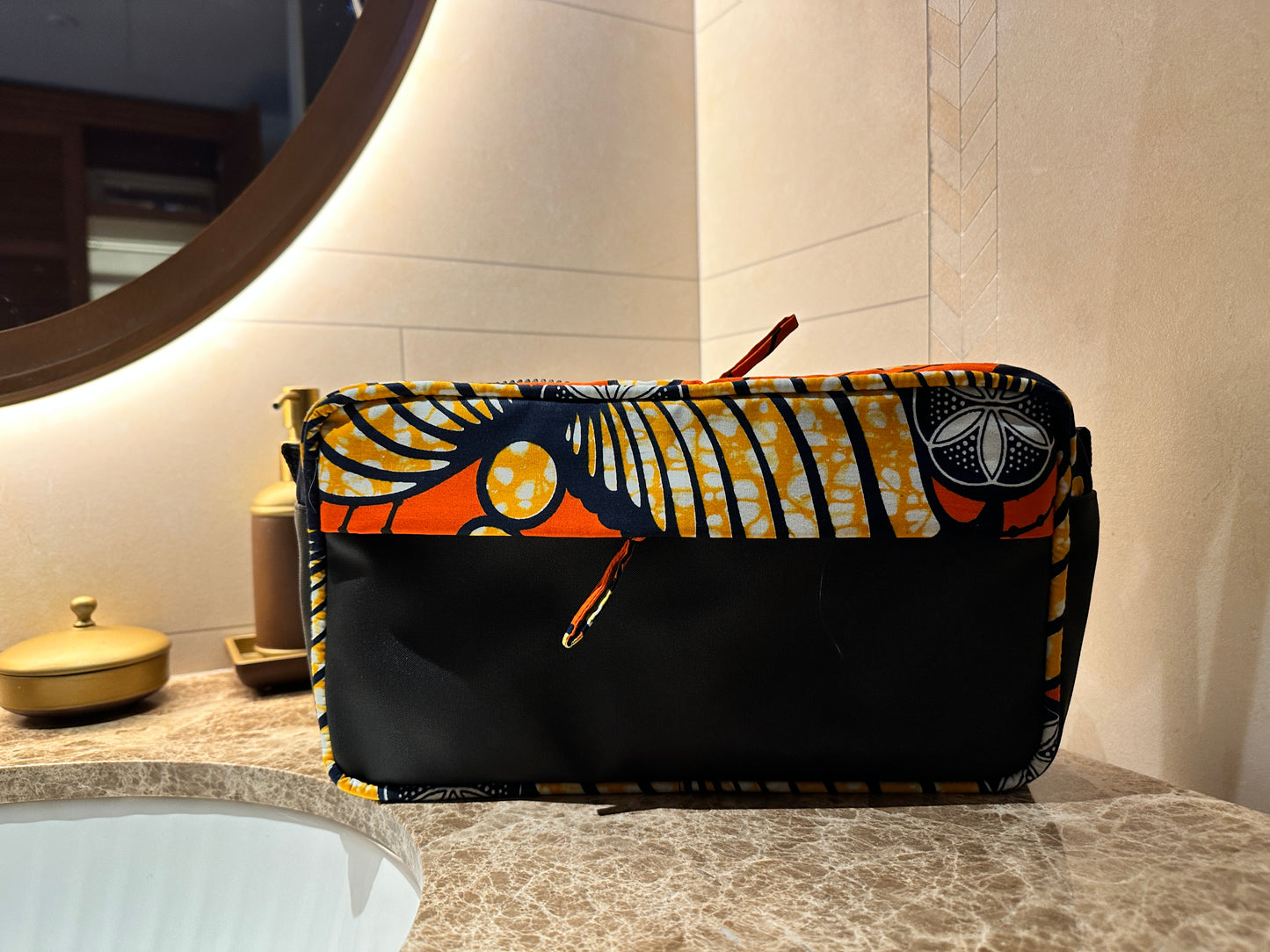 Afia Orange Sorbet African Print Wash Bag, located at the side of a handbasin and lit wall mirror and  metallic soap dish.