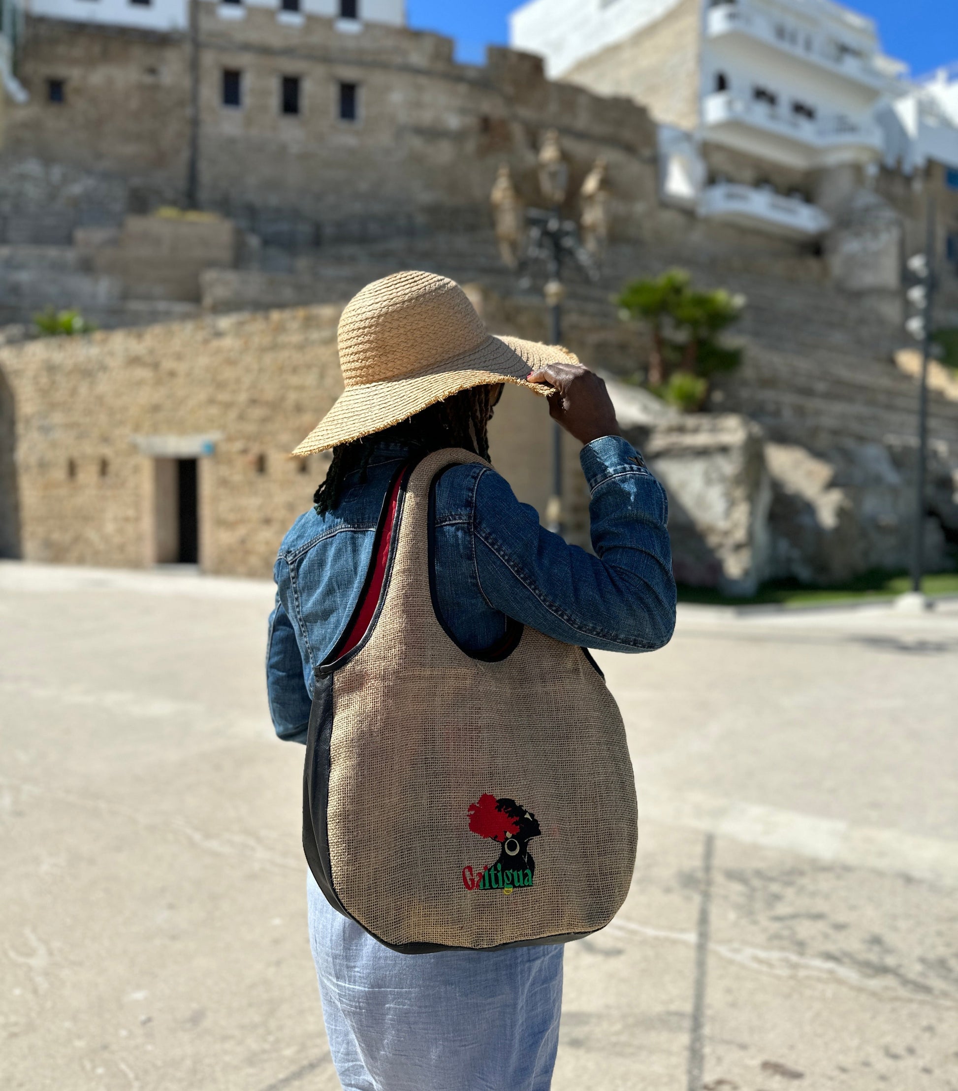 Woman with a white brim sun hat, wearing a blue denim jacket & dress wearing a Pan Africa Embroidered Galtigua logo, black leather & natural hessian design Kalia Hobo shoulder bag against a ruin castle in the back ground.