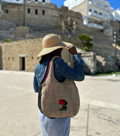 Woman with a white brim sun hat, wearing a blue denim jacket & dress wearing a Pan Africa Embroidered Galtigua logo, black leather & natural hessian design Kalia Hobo shoulder bag against a ruin castle in the back ground.