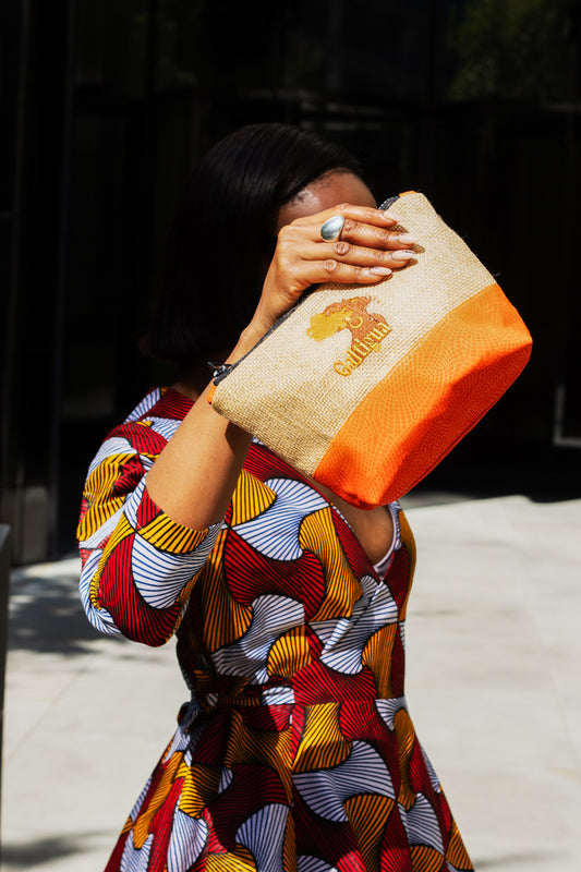 A young woman wearing an African Print blouse, holding Papaya orange and Hessian  Karissa pouch with galtigua embroidered logo.