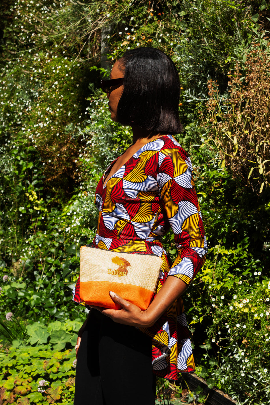 A young woman wearing an African Print skirt and black pencil skirt, holding an orange and Hessian cosmetic bag with galtigua embroidered logo.