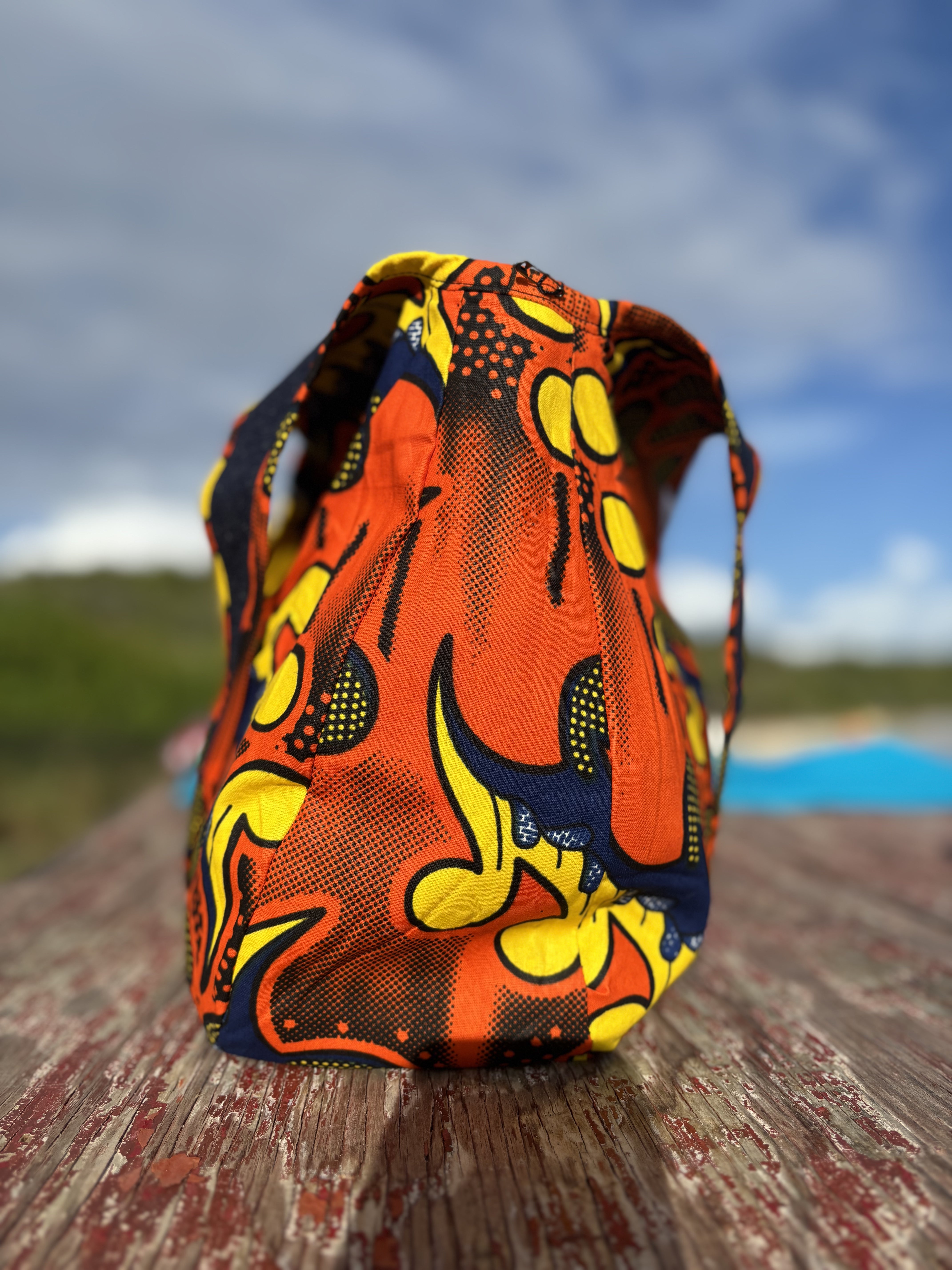 Reversible Denim citrus burst print Kalia Hobo shoulder bag, reserved to show the  Zumba African printed lining standing on a wooden bench against the back drop of the sea a cloudy sky and trees in the far distance.