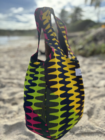 Reversible Denim Rock Candy print Kalia Hobo shoulder bag, reserved to show the  rock candy African printed lining standing balanced on a stick in the beach sand against the back drop of the sea with and palm trees in the far distance.