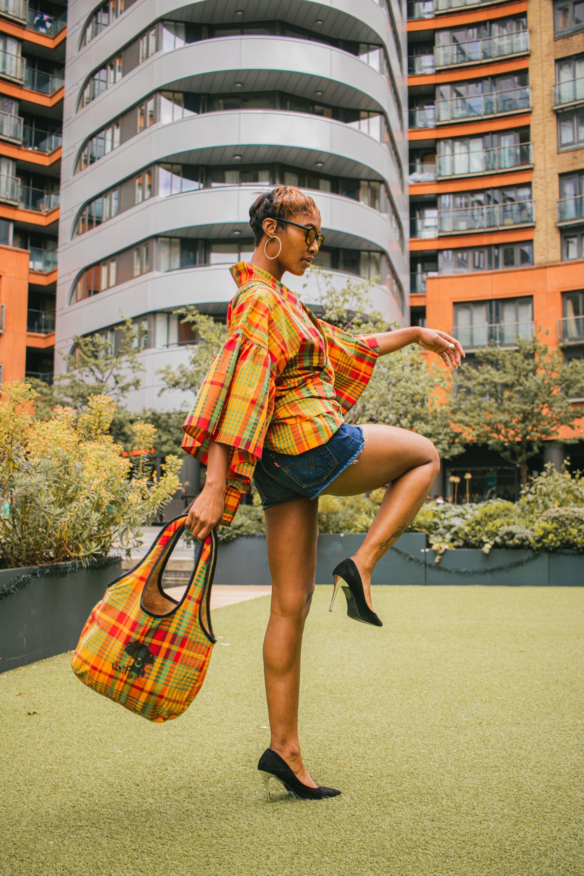 A  young woman wearing Caribbean Madras fabric top, shorts, black high heel shoes against the back drop of some foliage & sky rise buildings, standing swinging a denim Kalia Hobo shoulder bag with printed 'Galtigua'  black logo on it.