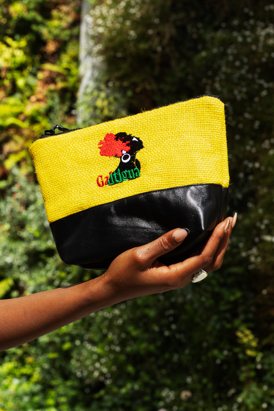 A young woman wearing a silver ring holding Soleil Yellow Hessian and black leather Karissa pouch with galtigua embroidered logo against a tree.